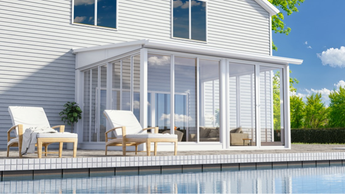 A glass sunroom with white framing attached to a house with white siding next to a pool deck with two lounge chairs next to a pool.